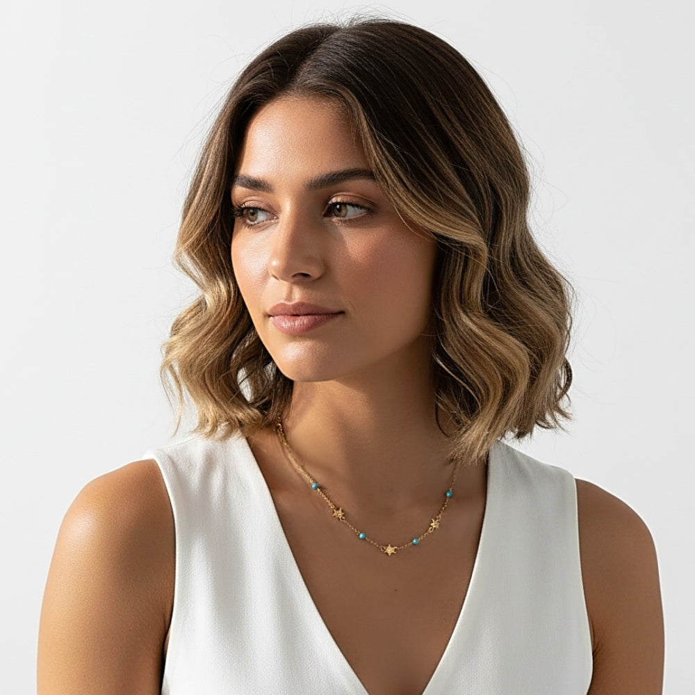 A woman with wavy brown and blonde hair wearing a delicate gold necklace with small star charms and turquoise blue beads, paired with a white sleeveless top against a light gray background.