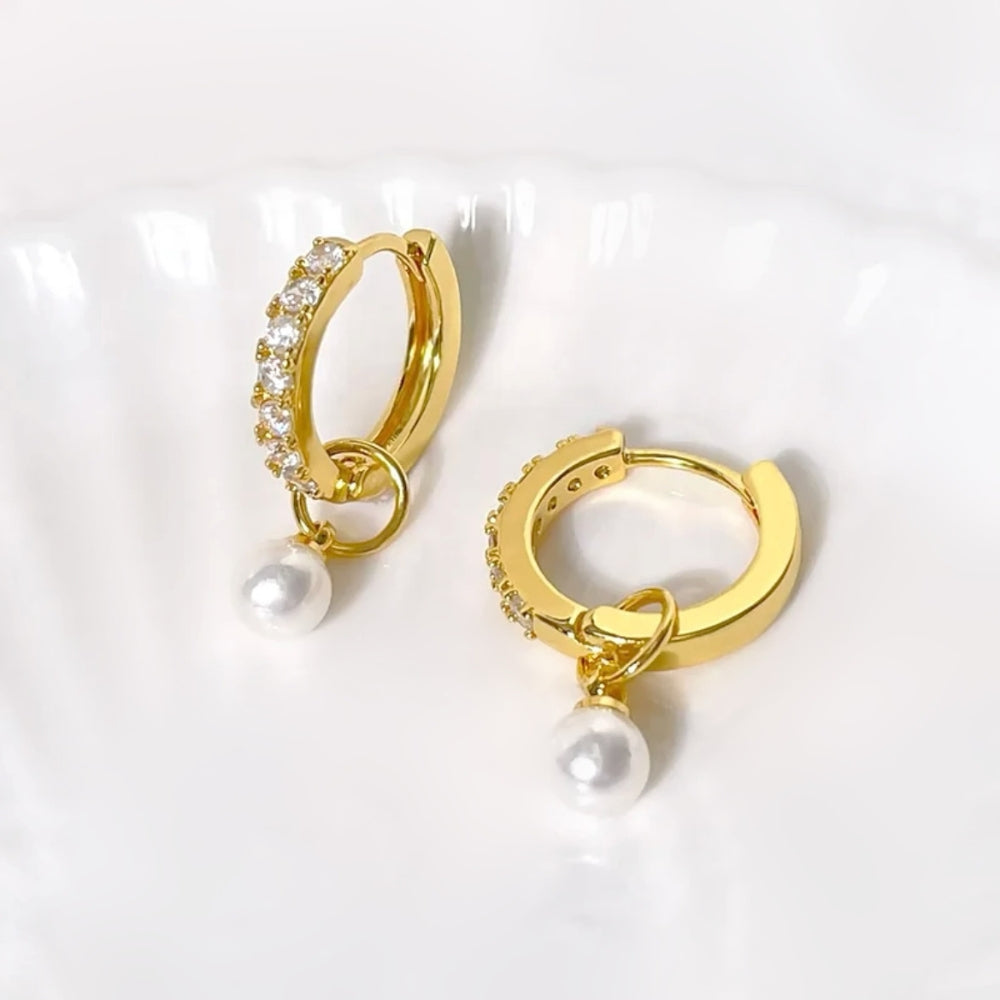 A pair of gold huggie hoop earrings with small clear stones along the front and a dangling white pearl, displayed on a white scalloped dish.