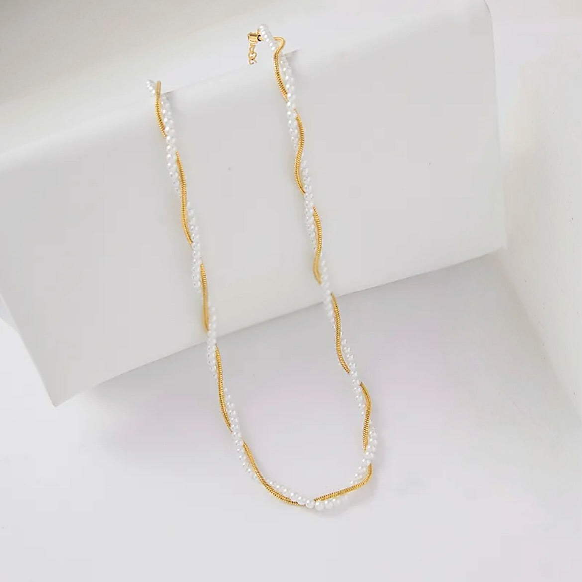 A delicate necklace featuring white freshwater pearls and a twisted gold-tone metal core, draped over a white display block.