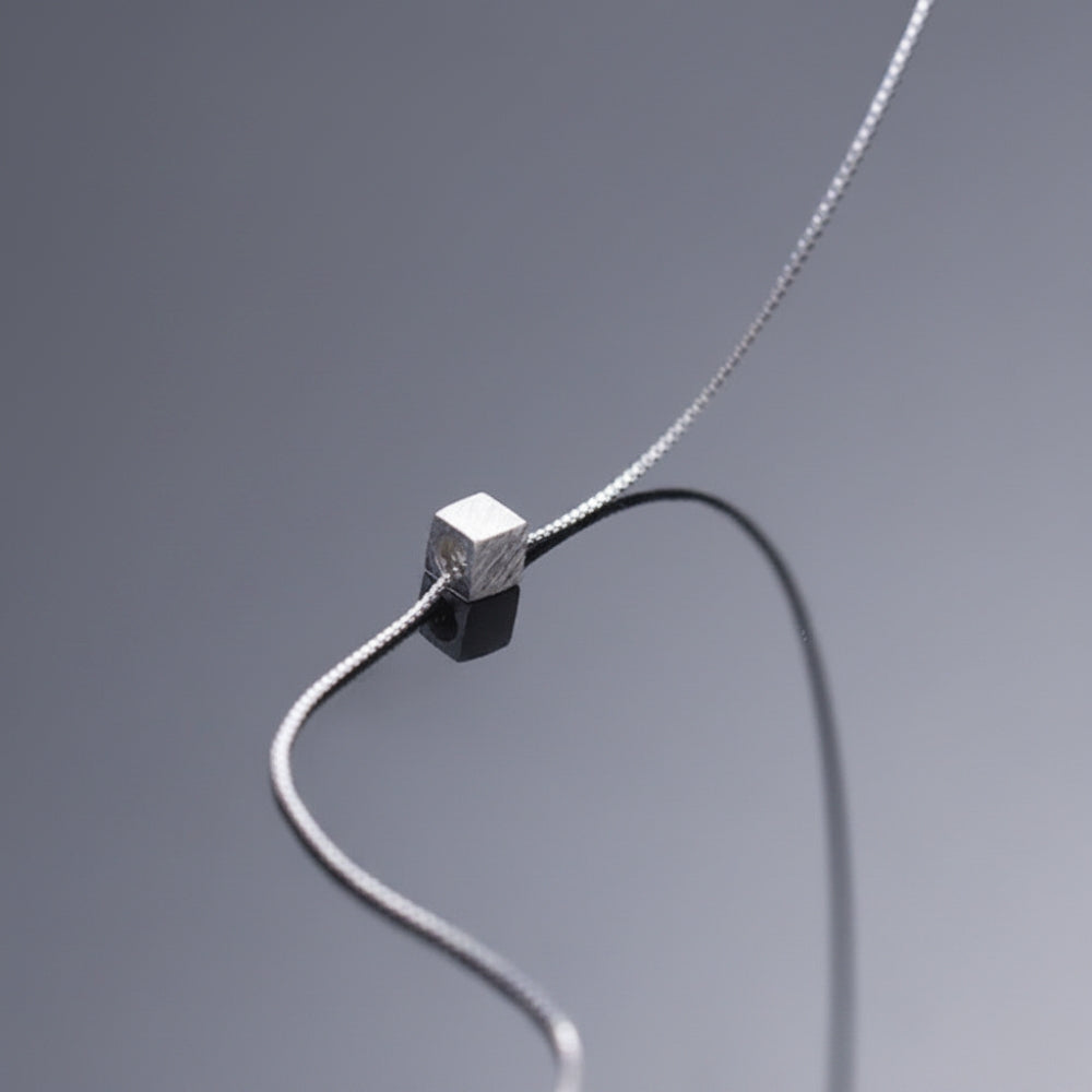 A silver chain necklace featuring a small, textured square cube bead, displayed on a reflective dark gray surface.