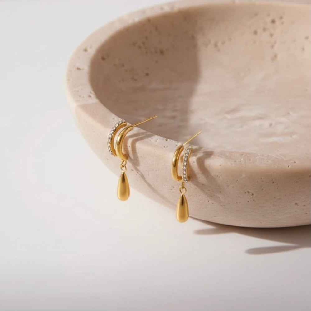 A pair of gold dangle earrings with small clear stone accents and teardrop-shaped drops, displayed on a beige textured ceramic dish against a white background.