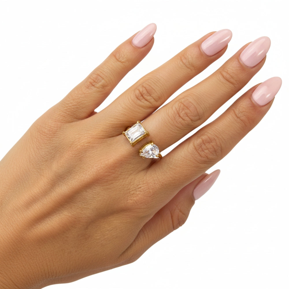 A hand with light pink manicured nails wearing two gold rings: one with a rectangular clear gemstone and one with a pear-cut clear gemstone, set against a plain white background.