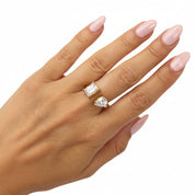 A hand with light pink manicured nails wearing two gold rings: one with a rectangular clear gemstone and one with a pear-cut clear gemstone, set against a plain white background.
