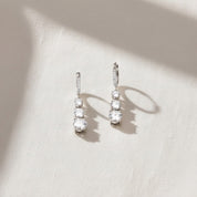 A pair of silver drop earrings with three clear oval-cut stones in each earring, resting on a textured off-white fabric surface with soft shadows.