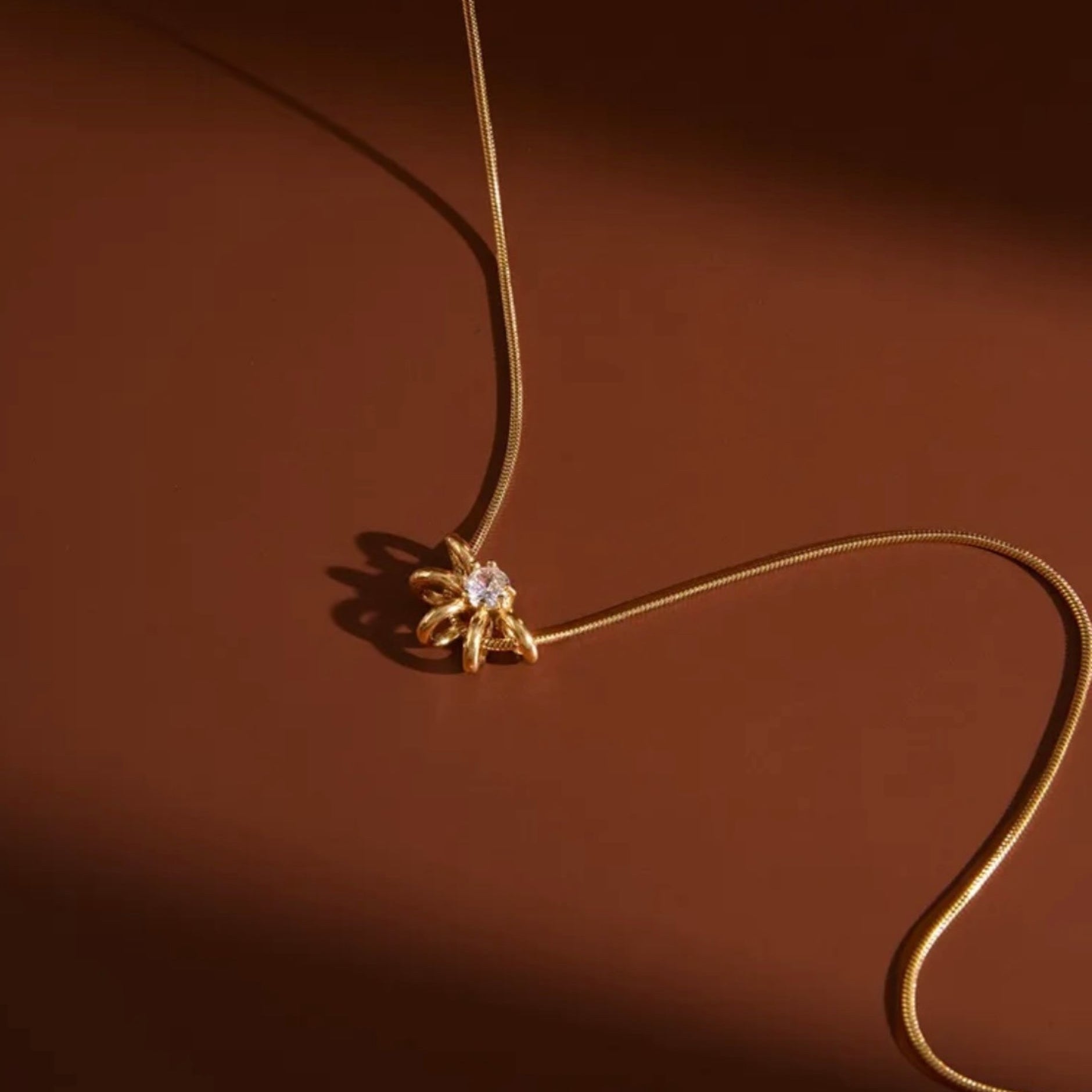 A delicate gold snake chain necklace with a small gold flower-like pendant set with a clear central stone, photographed on a warm brown background with dramatic shadows.