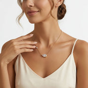 A woman wearing a delicate gold chain necklace with a teardrop-shaped clear gemstone pendant and a small matching stud earring, paired with a cream camisole top against a light gray background.