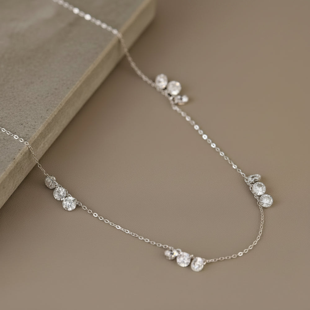 A delicate silver chain necklace with small clusters of clear round gemstones, displayed on a neutral beige surface next to a textured stone block.
