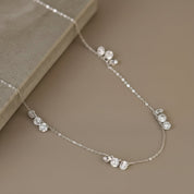 A delicate silver chain necklace with small clusters of clear round gemstones, displayed on a neutral beige surface next to a textured stone block.