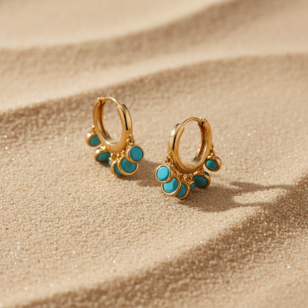 Gold huggie hoop earrings with small dangling turquoise stone clusters, photographed on a sandy beige surface in warm natural light.