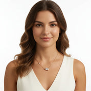 A woman with long brown hair wearing a delicate gold necklace with a clear teardrop-shaped crystal pendant and a small clear stone accent, paired with a white sleeveless top against a plain white background.