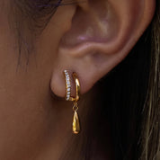 A close-up of a person's ear wearing a gold huggie hoop earring adorned with small clear stones and a dangling gold teardrop charm.