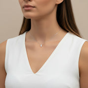 A close-up of a woman wearing a delicate silver chain necklace with a small pear-cut clear gemstone pendant, styled with a white sleeveless top against a neutral beige background.