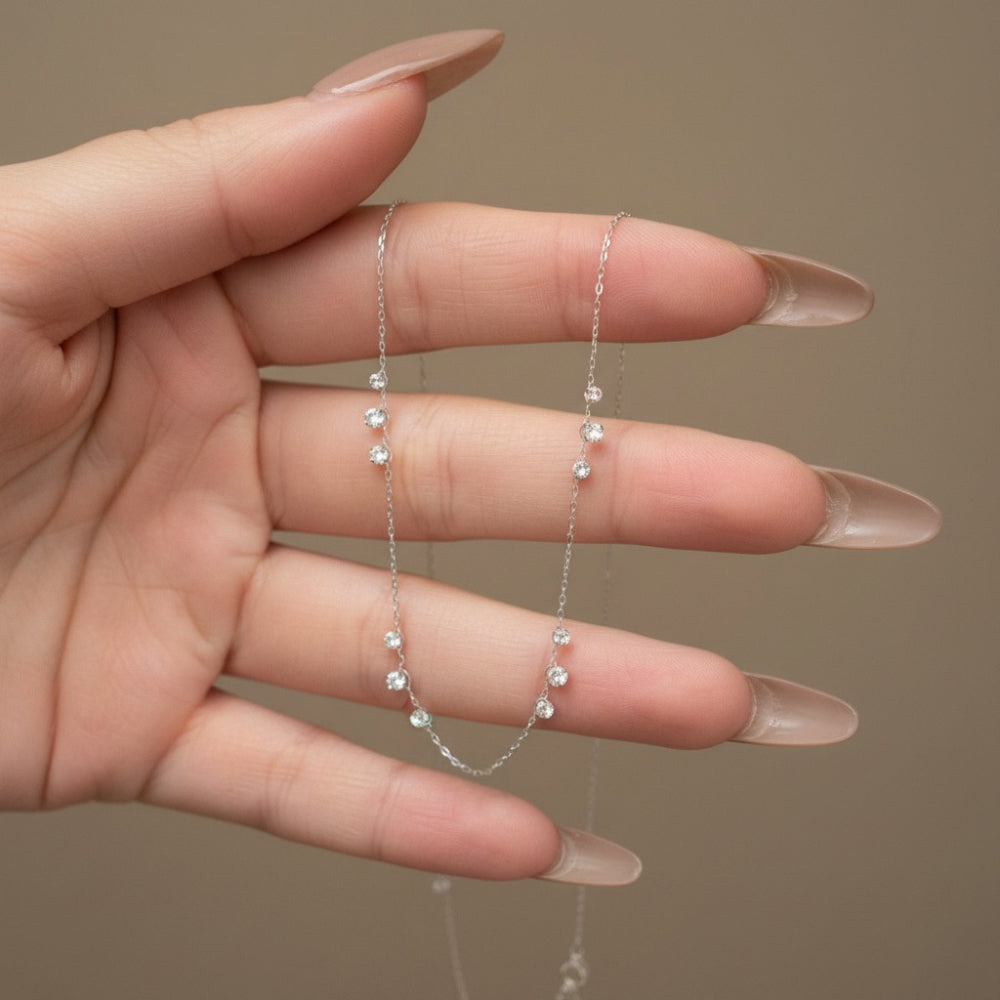 A delicate silver chain necklace with small clear crystal beads, held in a hand with long nude manicured nails against a solid taupe background.