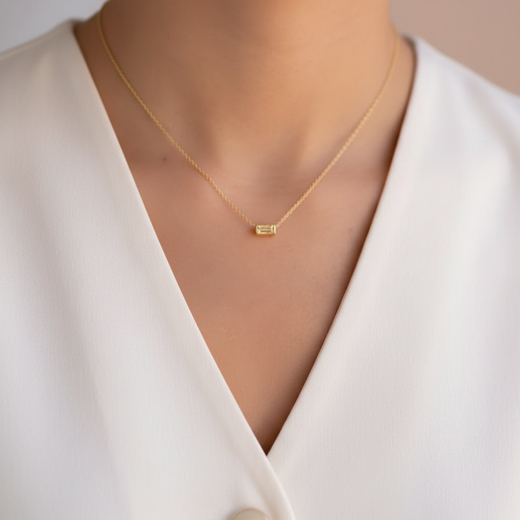 A delicate gold chain necklace with a small horizontal yellow pendant, worn by a model in a white V-neck top.