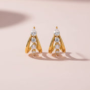A pair of gold geometric dangle earrings with clear marquise and round gemstones, displayed on a soft pink background.