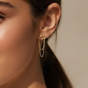 A close-up of a model's ear wearing a delicate gold double-hoop earring with two hanging chain drops, set against a warm beige background.