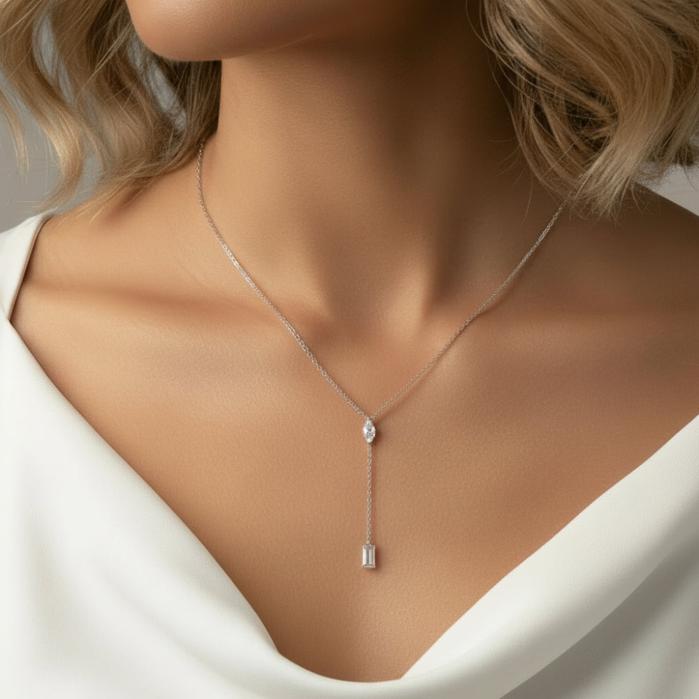 A close-up of a model wearing a delicate silver Y-necklace with a small teardrop-shaped clear gemstone and a matching rectangular clear stone pendant, paired with a white satin top.