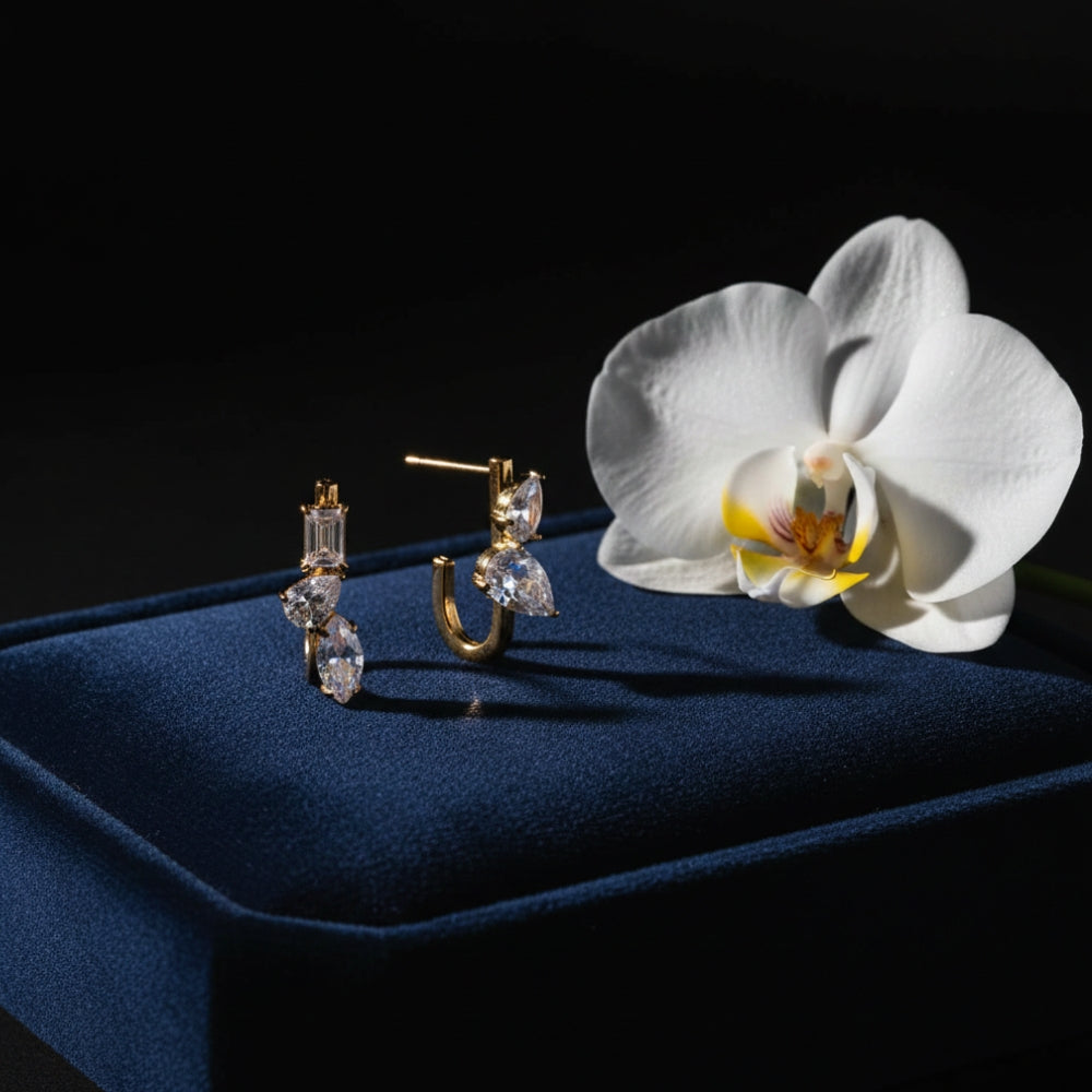 A pair of gold hoop earrings with clear marquise and oval-cut stones, displayed on a dark blue velvet jewelry box next to a white orchid flower against a black background.