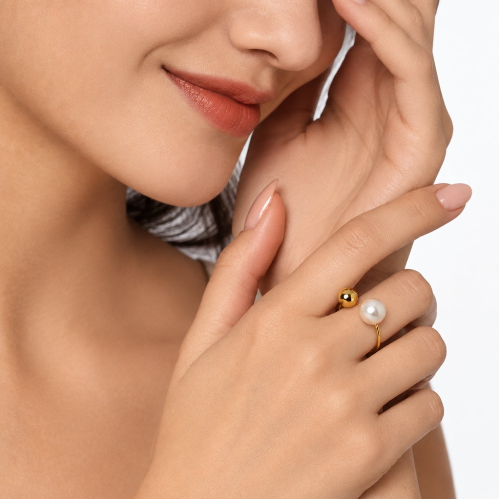 A close-up lifestyle shot of a model wearing a delicate gold ring with a small gold bead and a white pearl on a white background.