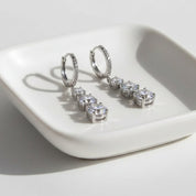 A pair of silver hoop earrings with four clear round-cut stones in a drop design, displayed on a white ceramic dish against a white background.