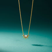 A delicate gold chain necklace with a small gold starburst pendant featuring a central clear gemstone, suspended against a dark teal background.
