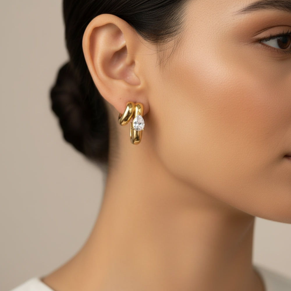 A close-up of a model's ear wearing a gold huggie hoop earring with a clear marquise-cut gemstone.