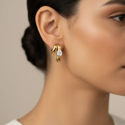 A close-up of a model's ear wearing a gold huggie hoop earring with a clear marquise-cut gemstone.