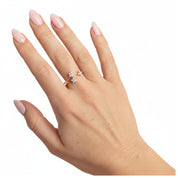 A close-up of a hand with pale pink manicured nails wearing a delicate gold ring with three small clear gemstones, set against a plain white background.