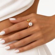 A close-up of a hand wearing a delicate gold ring with two pearl-like beads, one larger and lower, and one smaller, higher, set against a white top and light background.