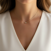 A close-up of a woman wearing a delicate gold chain necklace with a small rectangular bezel-set clear gemstone pendant, paired with a white V-neck top.