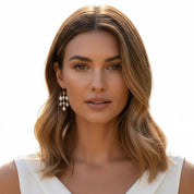 A woman with shoulder-length wavy brown hair wearing gold and pearl drop earrings, shown in a close-up portrait against a bright white background.