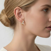 A close-up side profile of a woman with blonde hair in a bun, wearing a delicate gold hoop earring adorned with small clear gemstones, against a clean light gray background.
