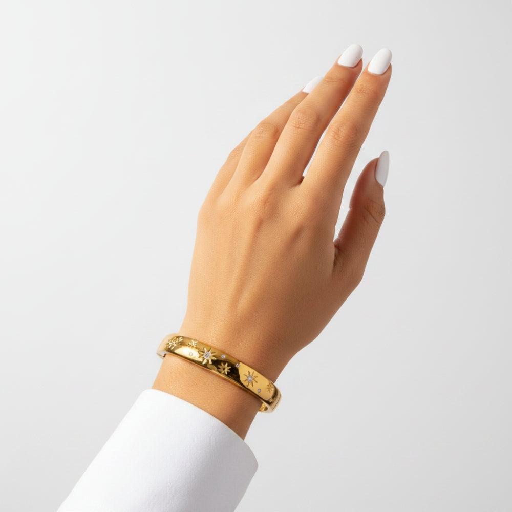 A gold bangle bracelet with engraved starburst motifs, shown on a model's wrist against a clean, light gray background.