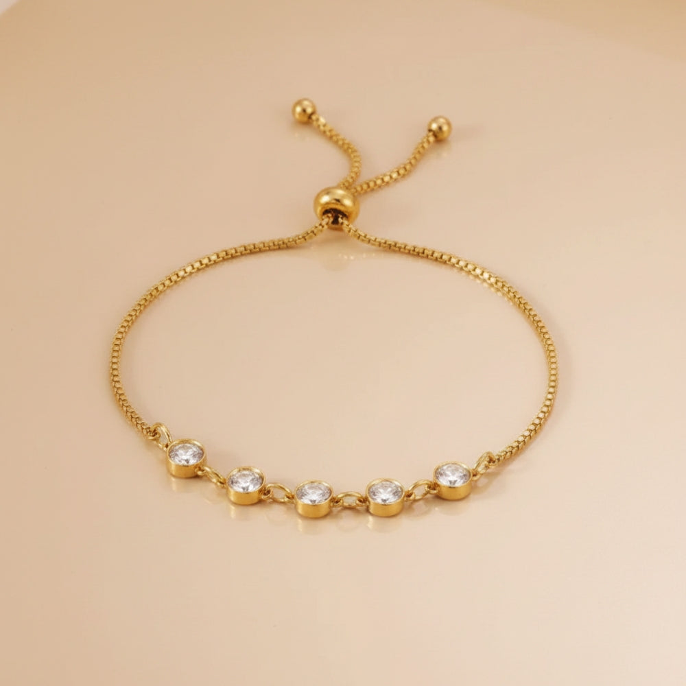 A delicate gold adjustable slide bracelet featuring five clear round-cut gemstones set in a row on the front, displayed on a warm beige background.
