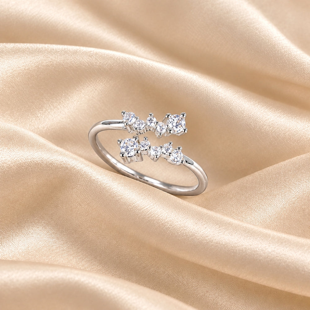 A silver ring with three small clear gemstones arranged in a bow-like cluster, photographed on draped champagne satin fabric.
