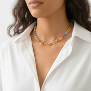 A close-up of a model wearing a colorful chain-link necklace with gold-tone metal links and translucent links in yellow, pink, blue, and orange, styled with a white collared shirt.
