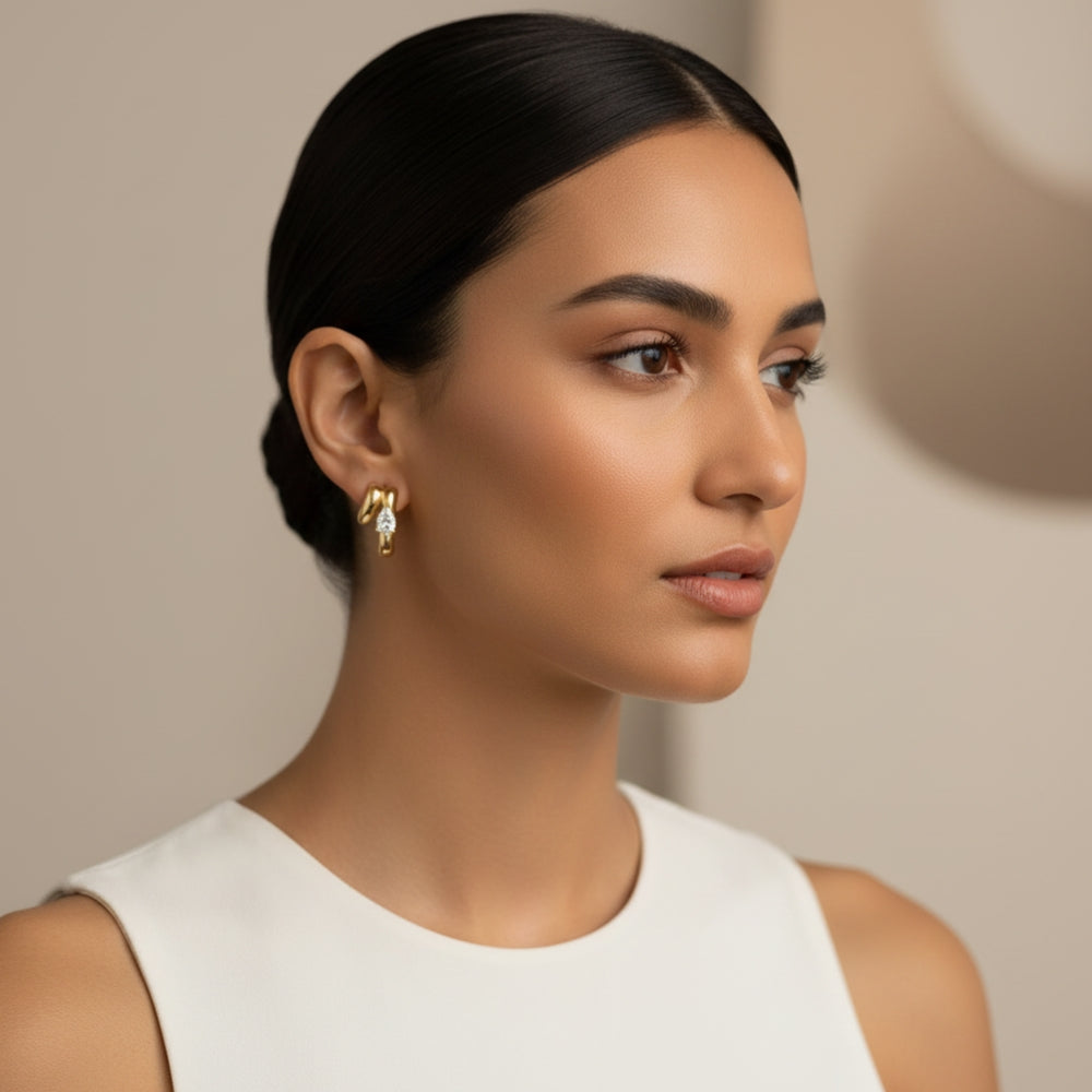 A close-up portrait of a woman with dark hair pulled back, wearing a small gold hoop earring with clear gemstones, against a soft beige background.