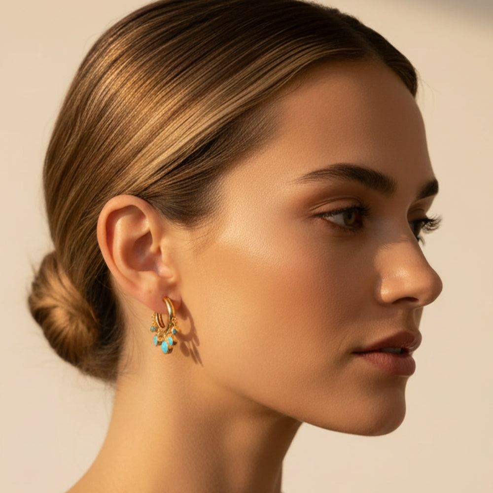 A close-up side profile of a woman with her hair in a low bun, wearing a delicate gold hoop earring adorned with small turquoise blue and yellow crystals, set against a warm beige background.