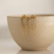 A pair of gold star-shaped dangle earrings with small clear stones at the top and layered chain drops, displayed on the rim of a speckled beige ceramic bowl against a soft neutral background.