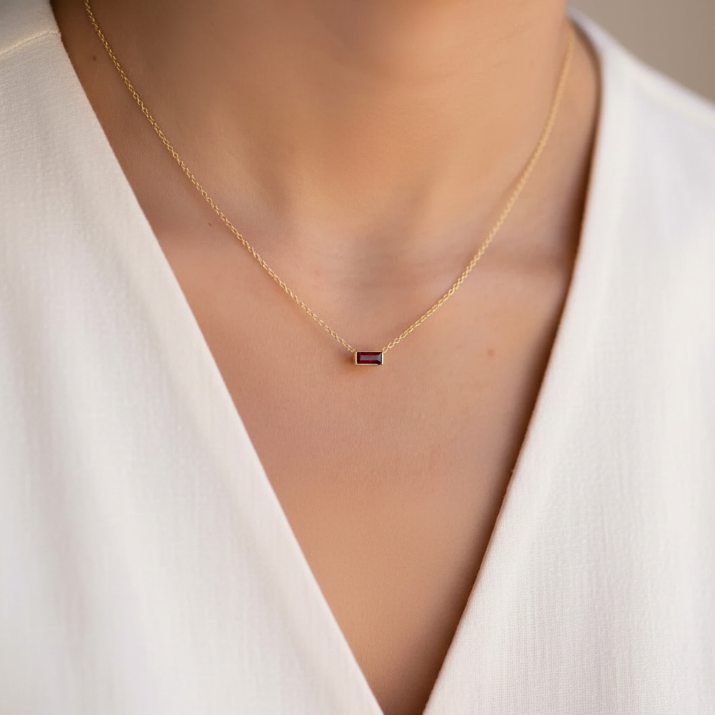 A delicate gold chain necklace with a small rectangular dark gemstone pendant, worn on a model's neck over a white V-neck top.