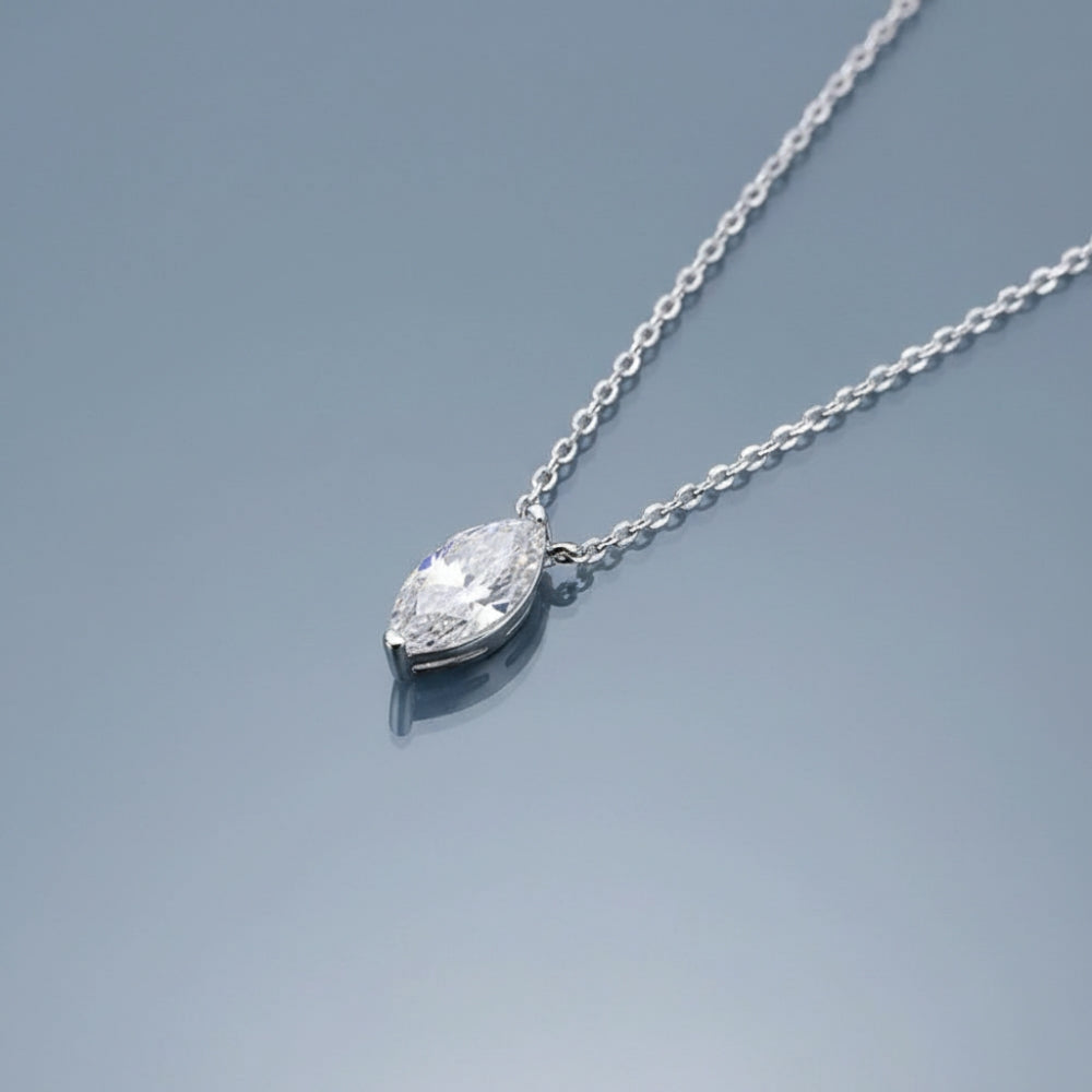 A silver necklace with a pear-cut clear gemstone pendant on a delicate silver chain, displayed on a reflective light blue surface.