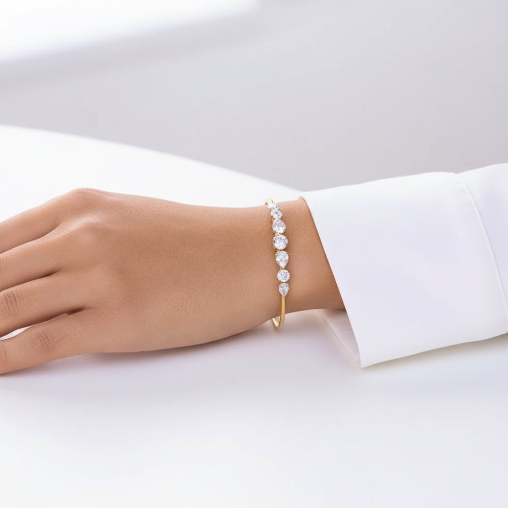 A delicate gold bangle bracelet with a row of sparkling clear gemstones, shown worn on a person's wrist with a white dress sleeve visible.