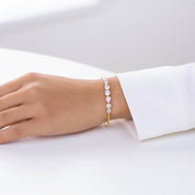 A delicate gold bangle bracelet with a row of sparkling clear gemstones, shown worn on a person's wrist with a white dress sleeve visible.
