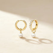 A pair of gold huggie hoop earrings adorned with small clear crystals and dangling white pearls, displayed on a warm beige surface with soft shadows.