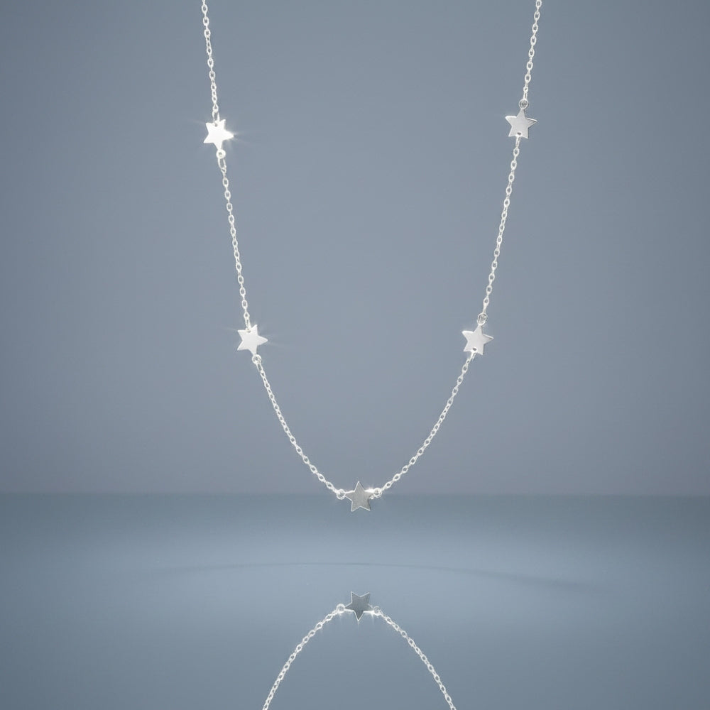 A delicate silver chain necklace featuring five small, evenly spaced star charms, displayed against a smooth, light blue-gray gradient background with a soft reflection below.