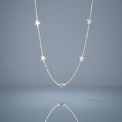 A delicate silver chain necklace featuring five small, evenly spaced star charms, displayed against a smooth, light blue-gray gradient background with a soft reflection below.