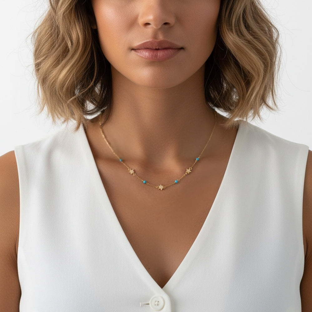 A close-up of a woman wearing a delicate gold necklace with small star and turquoise bead details, paired with a white sleeveless top.