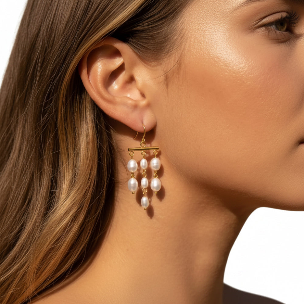 A close-up of a woman's ear wearing a gold dangle earring with three small white pearl drops.