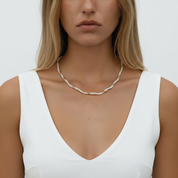 A close-up of a woman wearing a delicate white beaded necklace with small gold spacer beads, styled with a white sleeveless top against a light gray background.