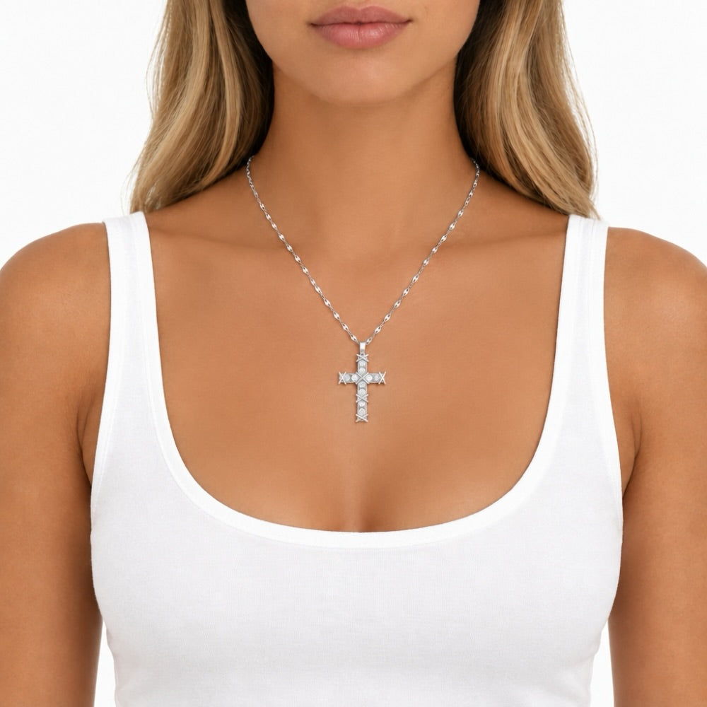 A close-up of a woman wearing a silver cross pendant necklace with clear stones, styled with a white ribbed tank top against a clean white background.